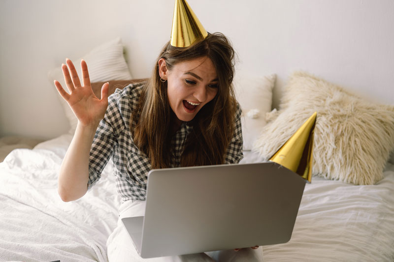 Mädchen hat ein Geburtstagshüttchen auf und feiert ihren Geburtstag online vor dem Laptop